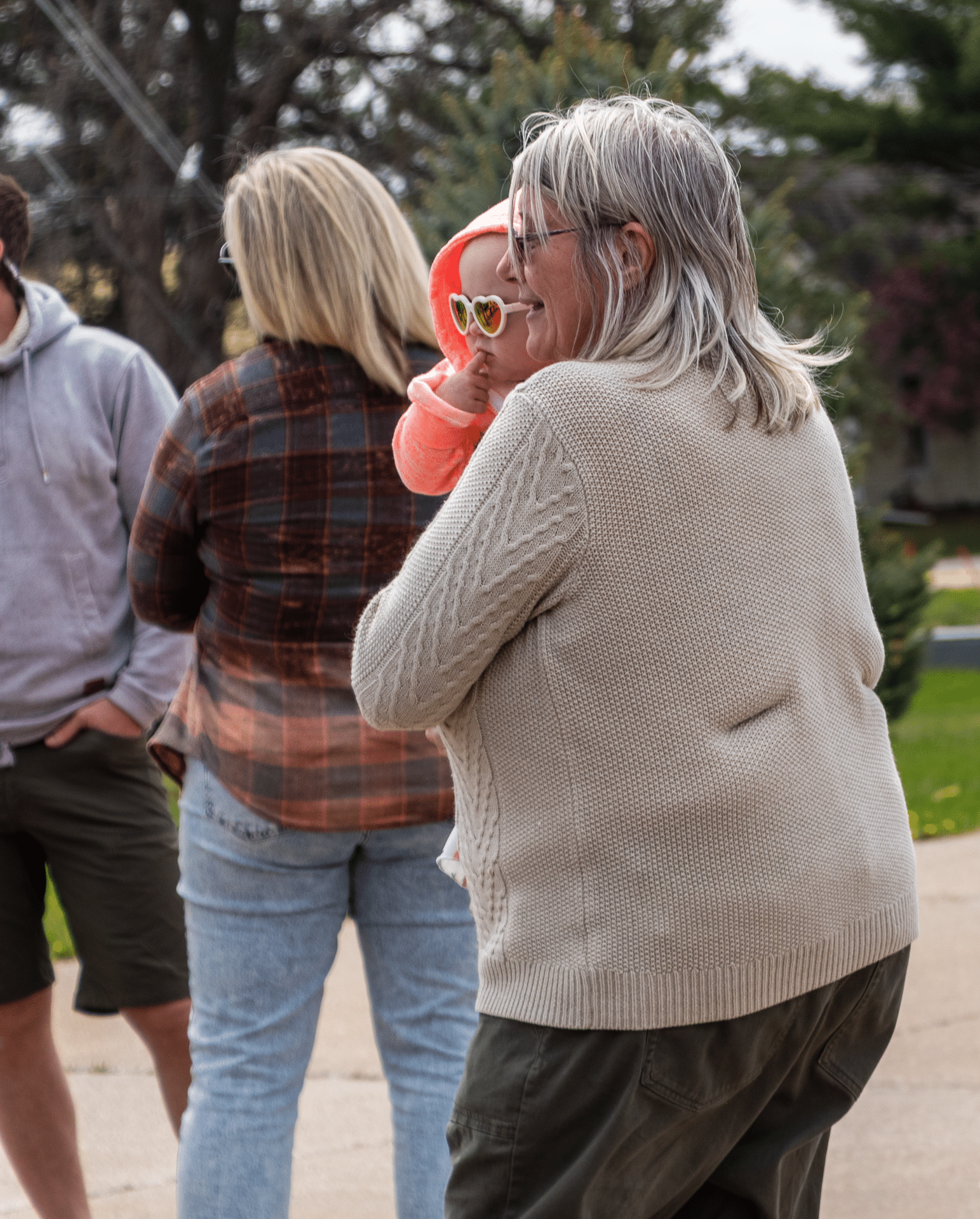 A caring woman holds a baby in sunglasses, smiling with friends—welcoming warmth and hope in our church family. - Muscatine Church of Christ