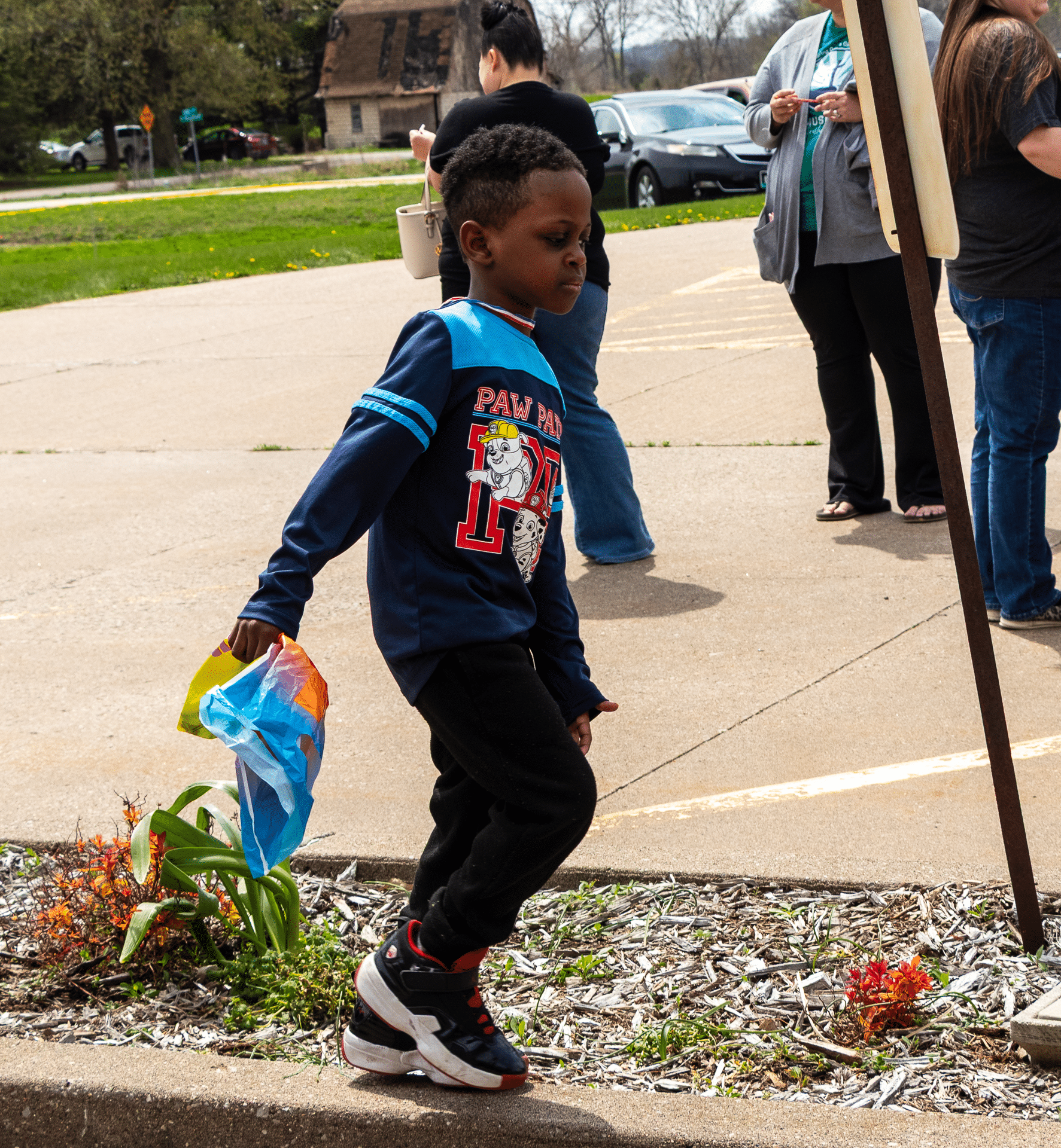A smiling boy with a bright bag walks near flowers, welcomed by caring adults at our church community gathering. - Muscatine Church of Christ
