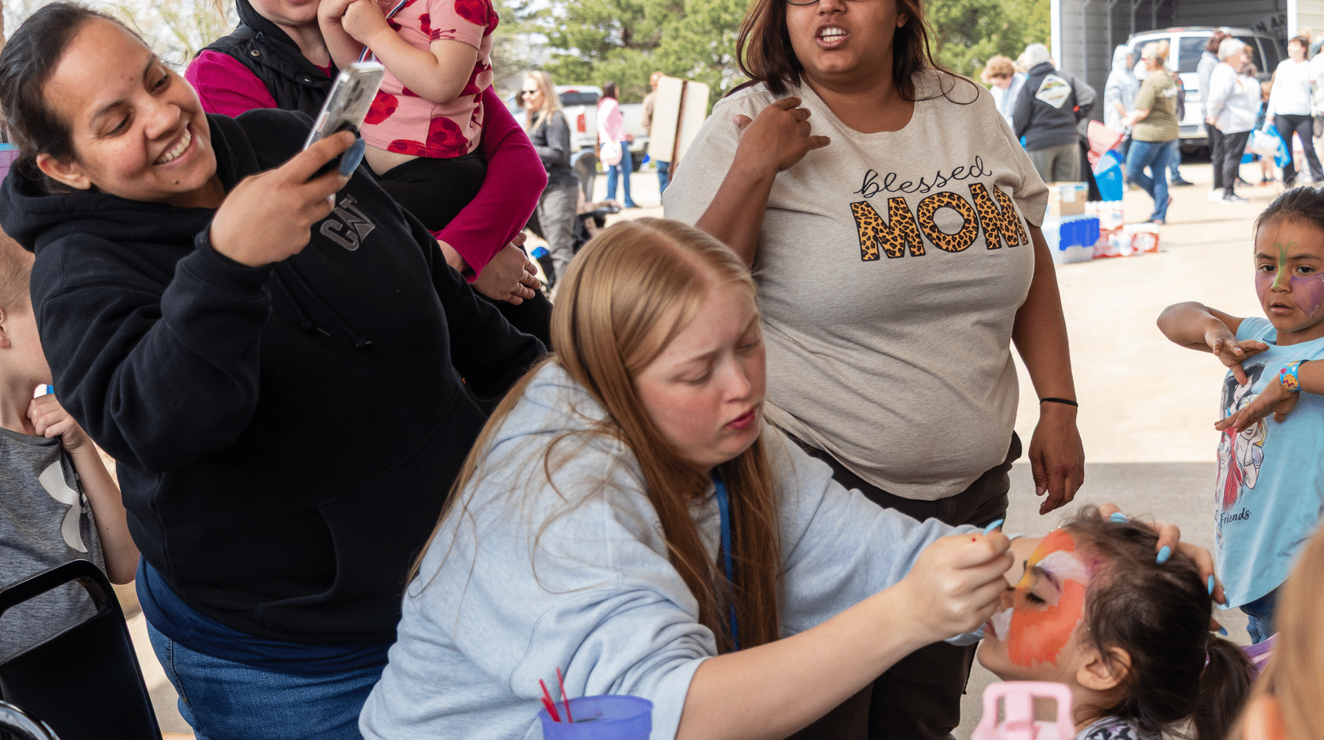 Smiling friends gather as a child’s face is painted, sharing joy and hope at our welcoming church event. - Muscatine Church of Christ