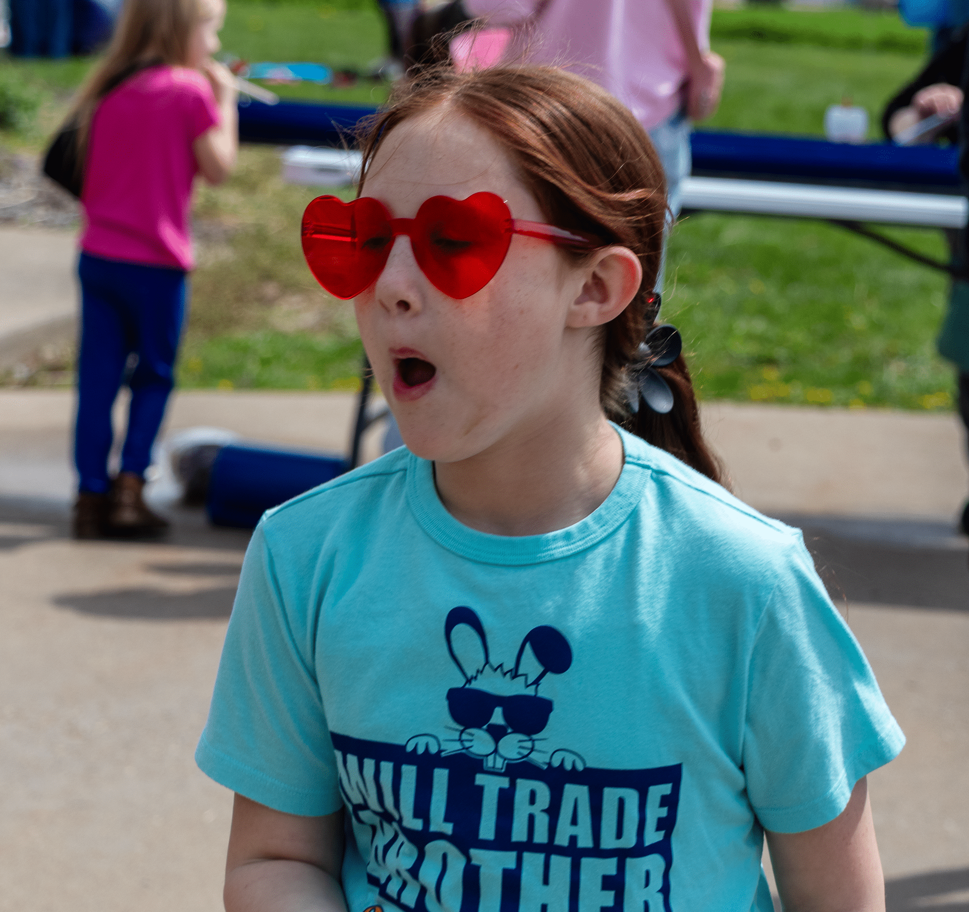 A joyful girl in red heart glasses smiles outdoors, surrounded by friends and welcome at our church gathering. - Muscatine Church of Christ