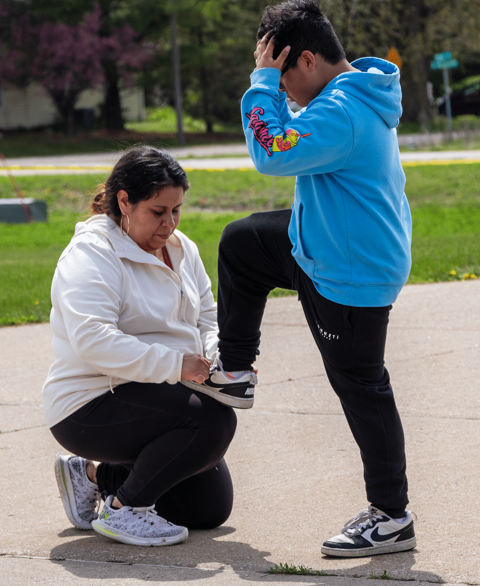A caring woman helps a boy tie his shoe, sharing kindness and support on the path—they are outdoors together. - Muscatine Church of Christ