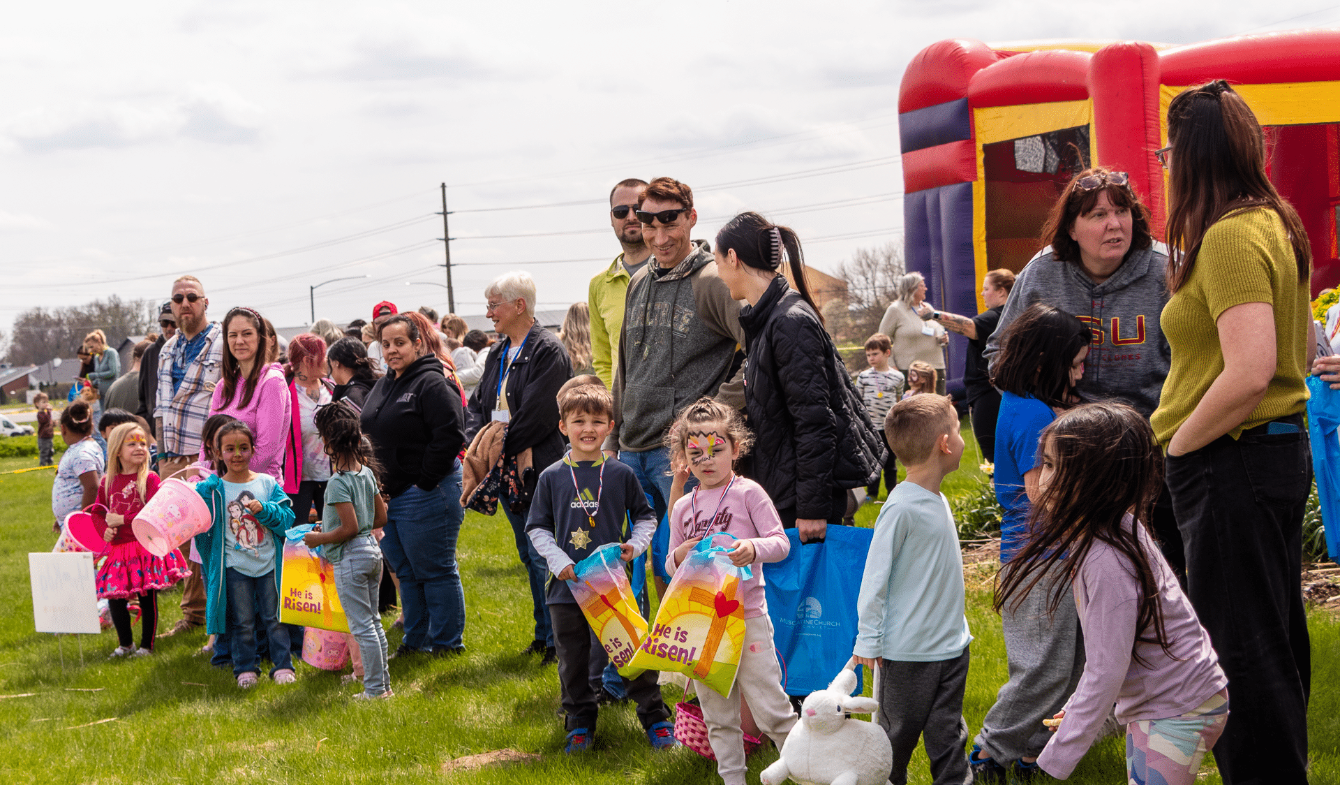 Children and adults gather with baskets, smiling together by a bounce house at our joyful outdoor church event. - Muscatine Church of Christ