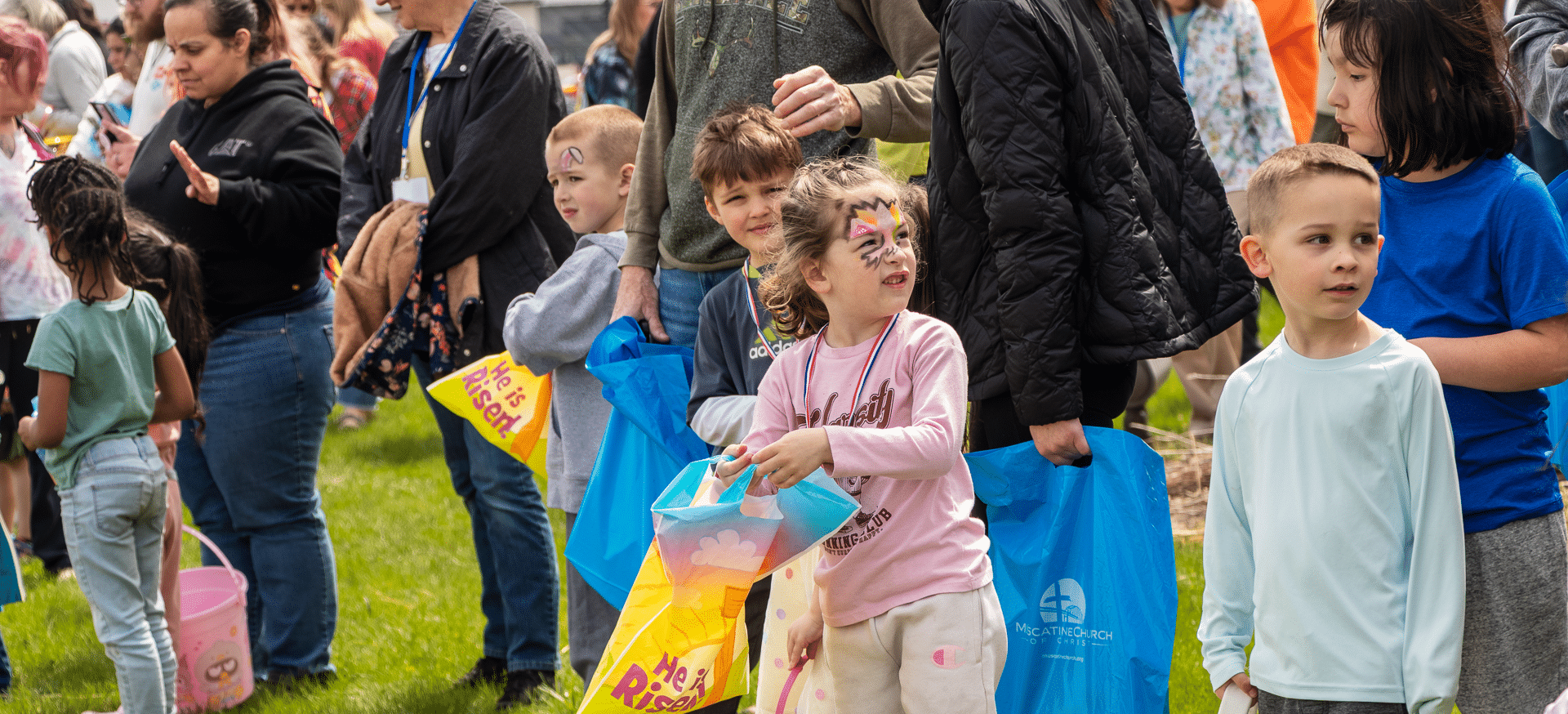 Smiling children with bags gather on the grass, surrounded by our joyful, welcoming church family. - Muscatine Church of Christ