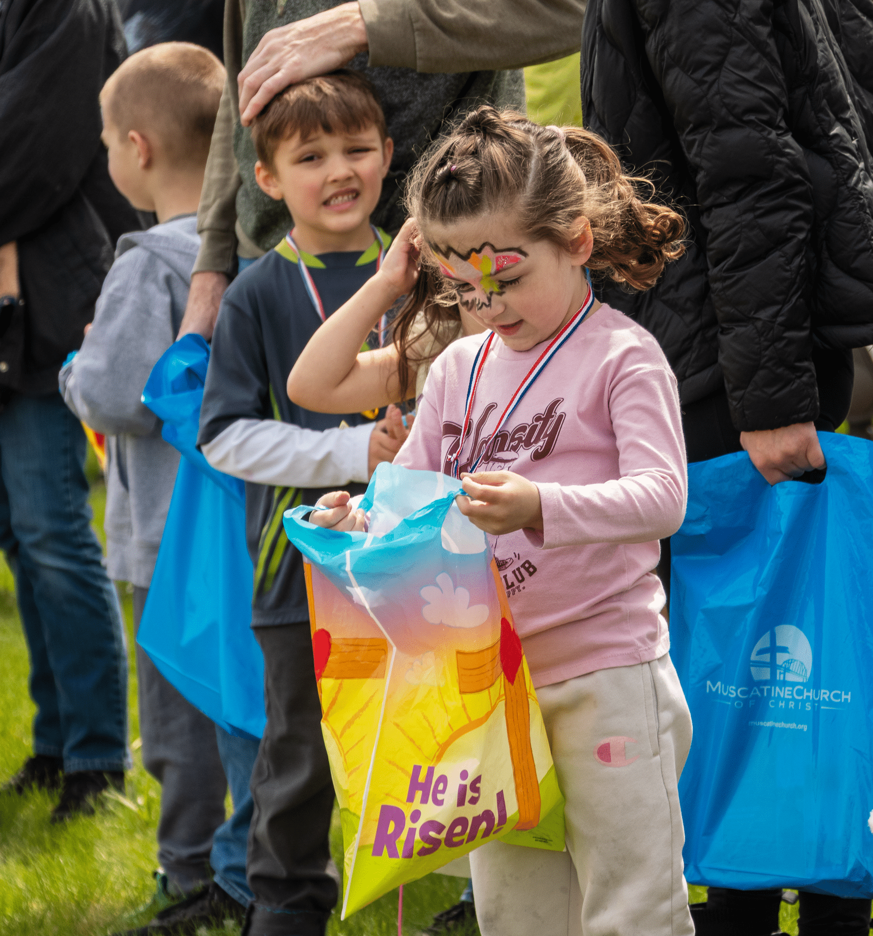 Smiling children with Easter bags reading He is Risen! gather outside, celebrating hope and community together. - Muscatine Church of Christ