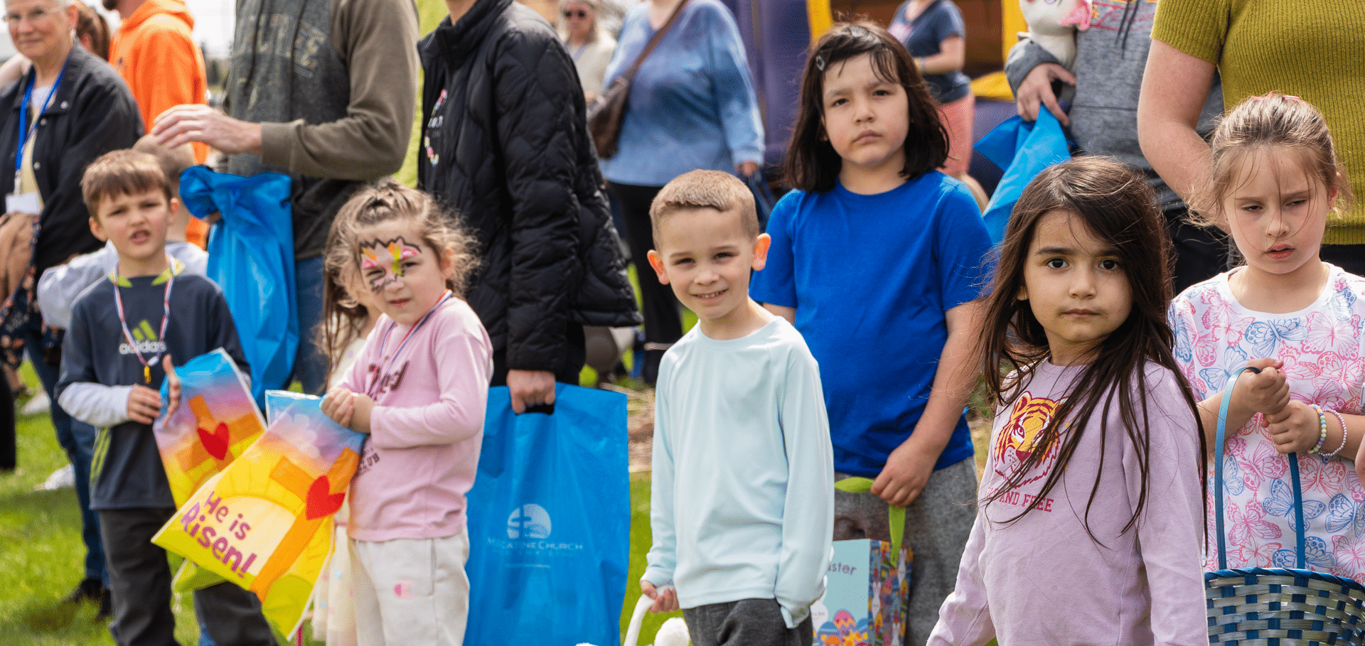 Smiling children with colorful bags gather on the grass, surrounded by caring adults and friends, sharing hope and joy. - Muscatine Church of Christ