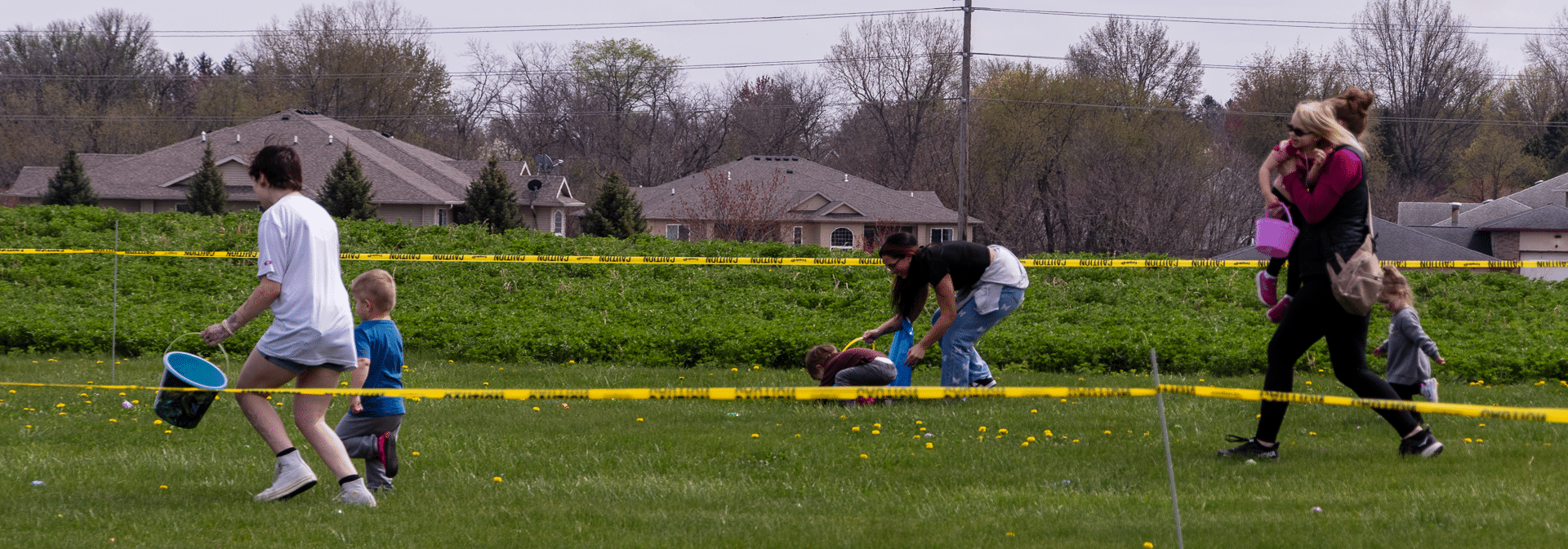 Adults and children share smiles and support on a sunny field, surrounded by welcoming homes and trees. - Muscatine Church of Christ