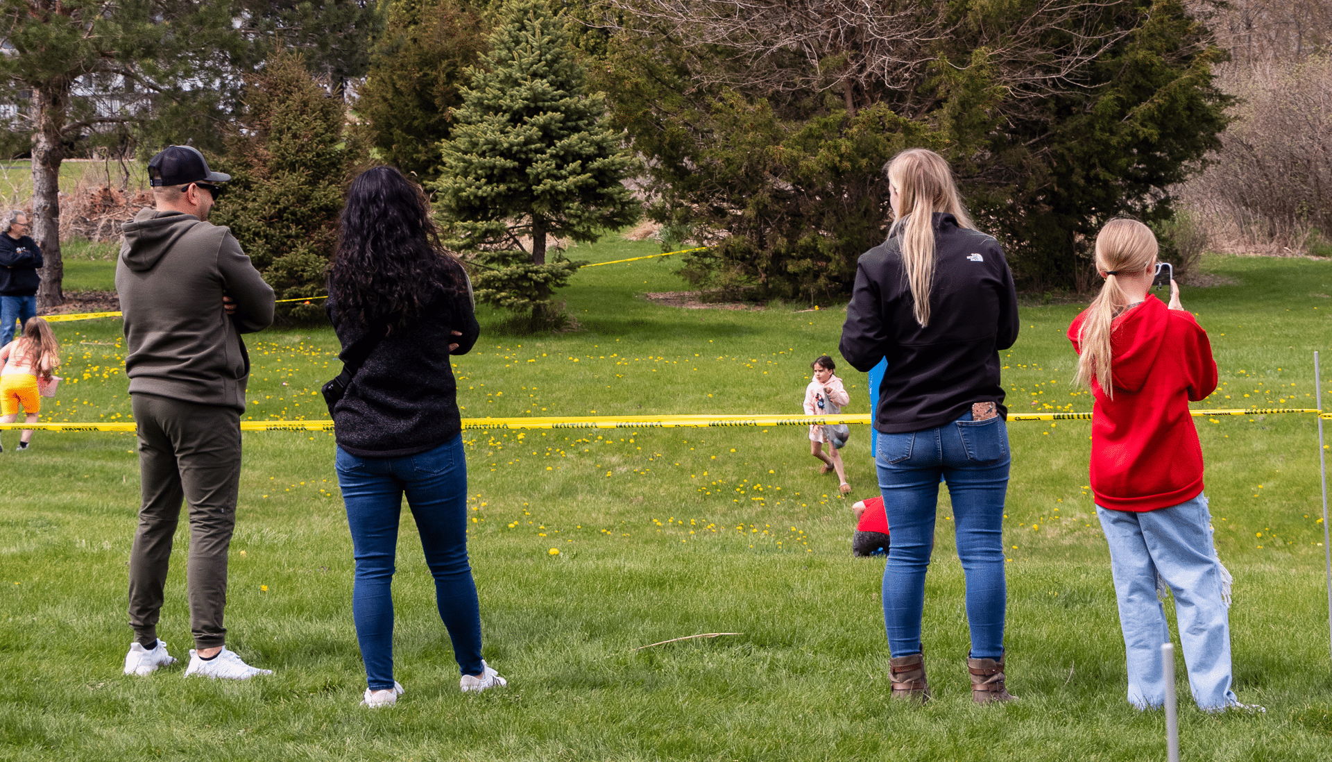 Four friends watch children play outdoors, with trees behind them—a joyful, welcoming moment in our church community. - Muscatine Church of Christ