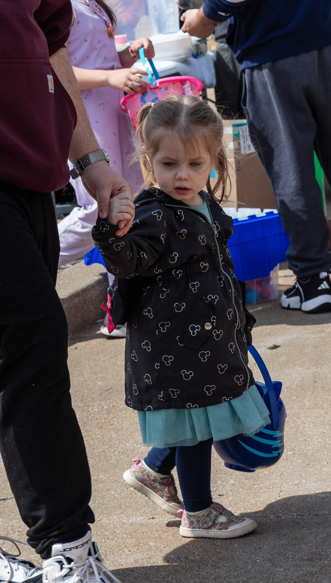 A young girl warmly holds an adult’s hand, carrying a blue basket, surrounded by our caring church family outdoors. - Muscatine Church of Christ