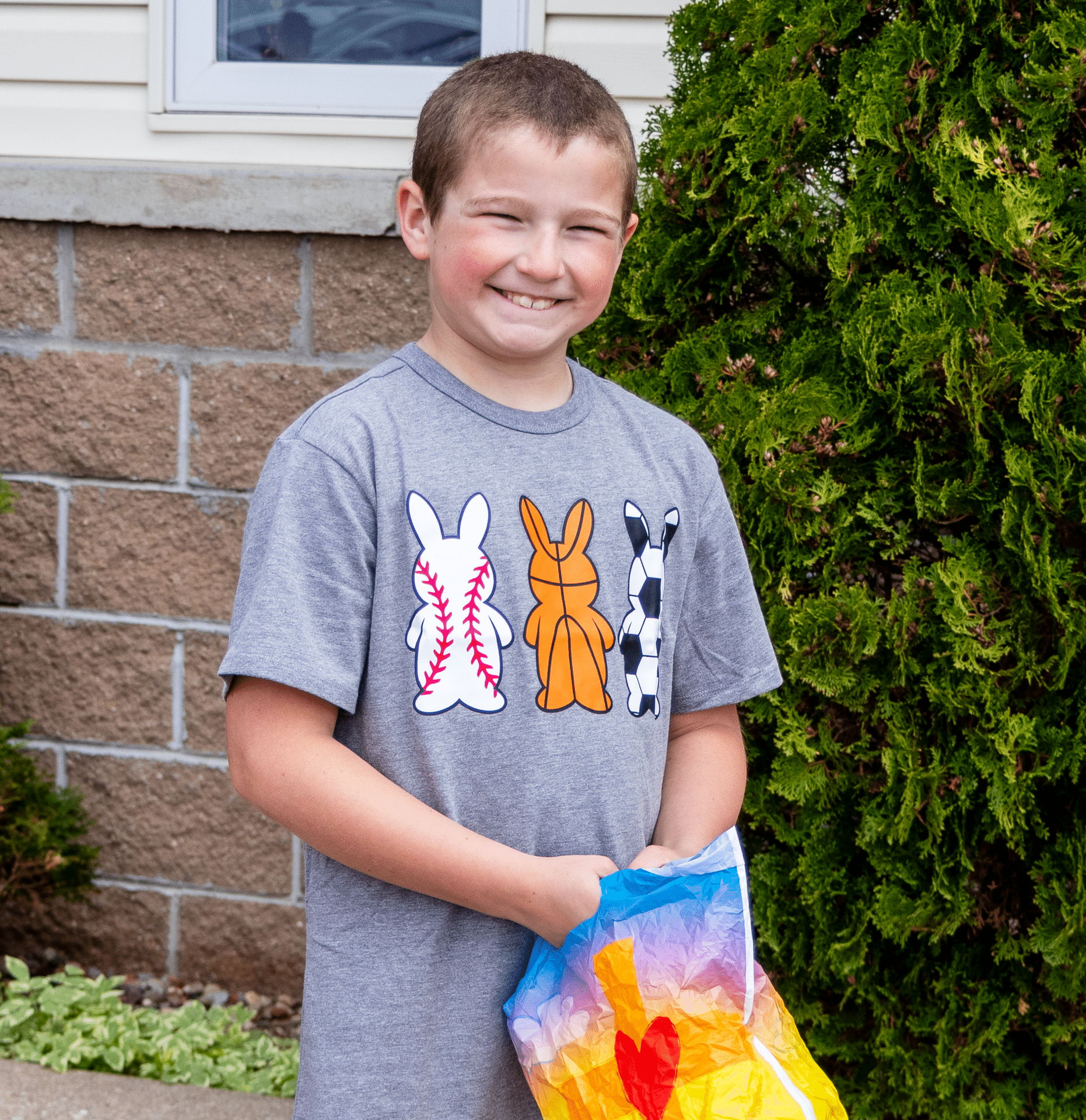 Smiling boy outdoors with a colorful bag, wearing a playful bunny sports shirt—celebrating joy and community together. - Muscatine Church of Christ