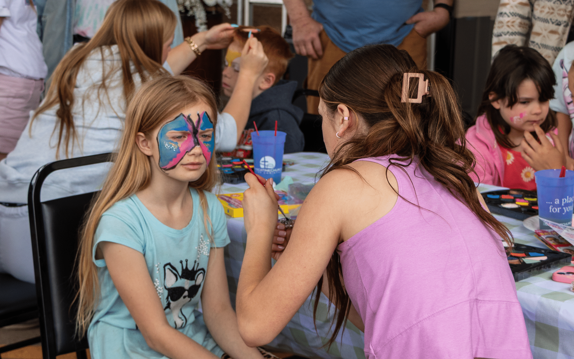 Smiling children gather around a table, sharing laughter, face painting, and friendship at church. All are welcome!. - Muscatine Church of Christ