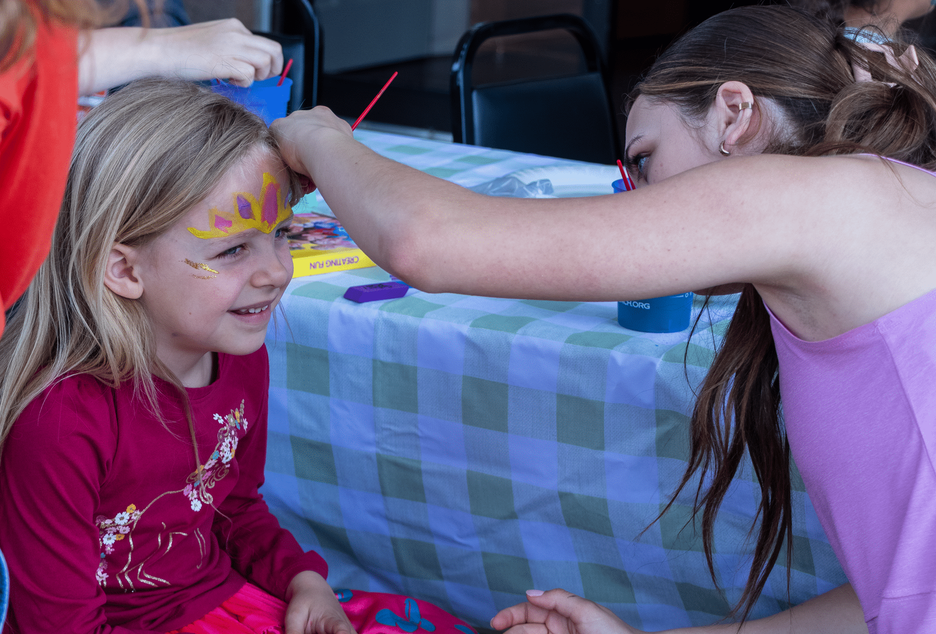 A smiling girl gets a sun painted on her forehead, sharing joy and hope at our welcoming church community table. - Muscatine Church of Christ