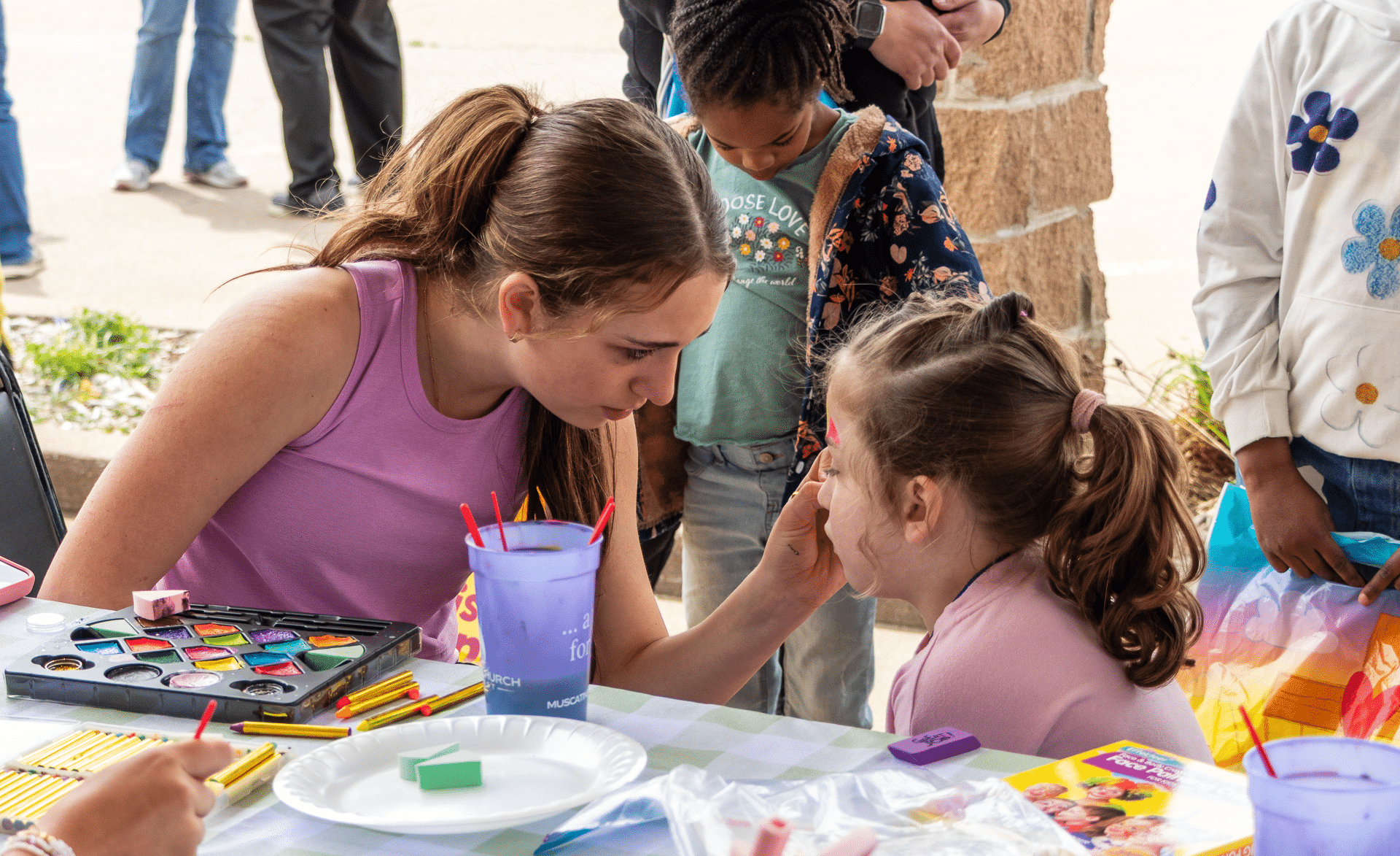A friendly volunteer paints a child’s face at our outdoor event, as children gather and laughter fills the air. - Muscatine Church of Christ
